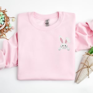 May include: A pink sweatshirt with a white bunny peeking out from the pocket. The bunny has pink ears and a black nose.