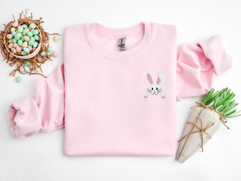 May include: A pink sweatshirt with a white bunny peeking out from the pocket. The bunny has pink ears and a black nose.