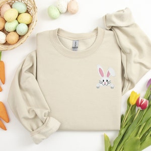 May include: A beige sweatshirt with a white bunny rabbit peeking out from the pocket. The bunny has pink ears and a black nose. The sweatshirt is surrounded by Easter decorations, including a basket of colorful eggs, carrots, and tulips.