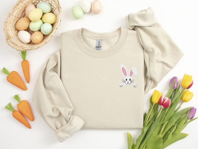 May include: A beige sweatshirt with a white bunny rabbit peeking out from the pocket. The bunny has pink ears and a black nose. The sweatshirt is surrounded by Easter decorations, including a basket of colorful eggs, carrots, and tulips.
