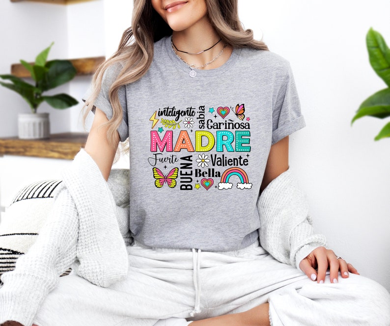 May include: A gray t-shirt with a colorful graphic design that says "MADRE" surrounded by words in Spanish, including "inteligente", "sabia", "cari&ntilde;osa", "fuerte", "buena", "valiente", and "bella".