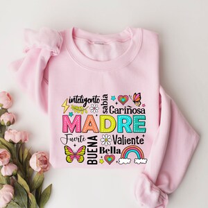 May include: Pink sweatshirt with a colorful graphic design that says "MADRE" surrounded by words in Spanish, including "inteligente", "sabia", "cari&ntilde;osa", "fuerte", "valiente", "buena", and "bella".