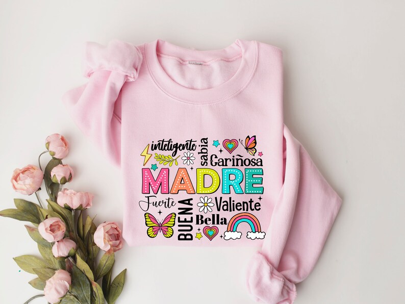 May include: Pink sweatshirt with a colorful graphic design that says "MADRE" surrounded by words in Spanish, including "inteligente", "sabia", "cari&ntilde;osa", "fuerte", "valiente", "buena", and "bella".