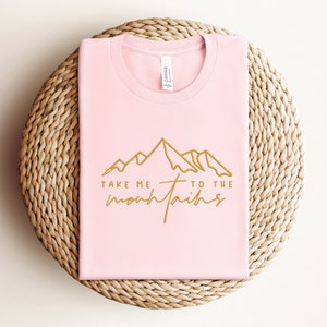 May include: Pink t-shirt with a gold outline of mountains and the text "Take me to the mountains".