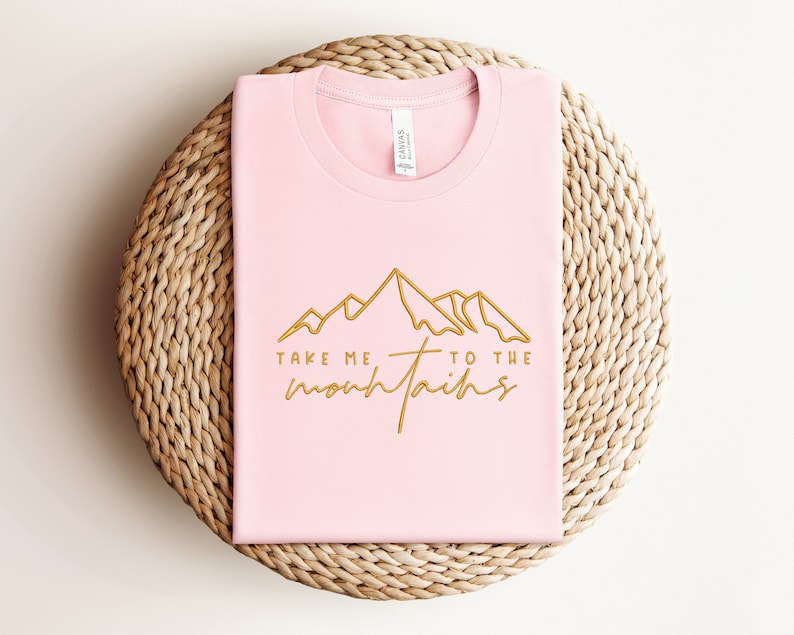 May include: Pink t-shirt with a gold outline of mountains and the text "Take me to the mountains".