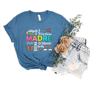May include: A teal blue t-shirt with a colorful graphic design that says "MADRE" surrounded by words in Spanish, including "inteligente", "sabia", "cari&ntilde;osa", "fuerte", "valiente", and "bella".