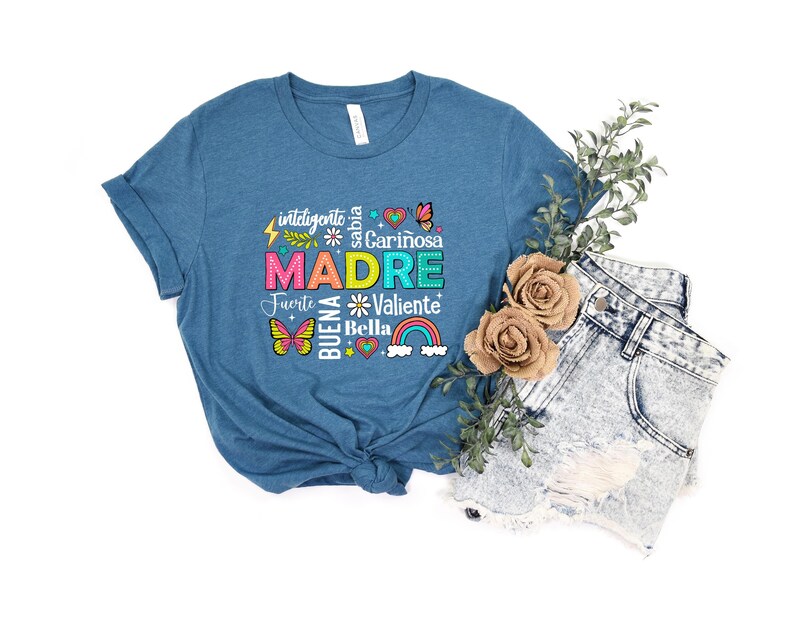 May include: A teal blue t-shirt with a colorful graphic design that says "MADRE" surrounded by words in Spanish, including "inteligente", "sabia", "cari&ntilde;osa", "fuerte", "valiente", and "bella".