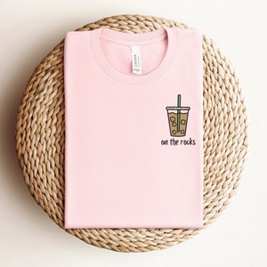 K&ouml;nnte beinhalten: Ein pinkfarbenes T-Shirt mit einer Grafik eines Glases mit Eiskaffee und dem Text "on the rocks".