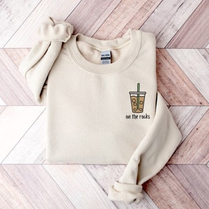 K&ouml;nnte beinhalten: Ein beiger Pullover mit einem Aufdruck eines braunen Eiskaffees mit gr&uuml;nem Strohhalm und dem Text "on the rocks" auf der Vorderseite.
