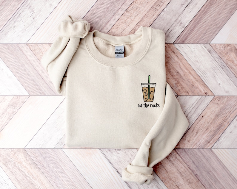 K&ouml;nnte beinhalten: Ein beiger Pullover mit einem Aufdruck eines braunen Eiskaffees mit gr&uuml;nem Strohhalm und dem Text "on the rocks" auf der Vorderseite.