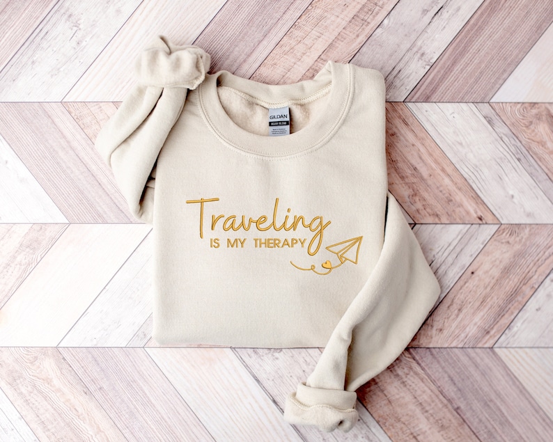 Może przedstawiać: Beżowa bluza z napisem "Traveling is my therapy" złotymi literami i grafiką papierowego samolotu.