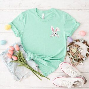May include: A light green t-shirt with a white bunny rabbit peeking out from the pocket. The shirt is laid flat on a white wooden surface with a pair of white sneakers, a bouquet of pink tulips, and Easter decorations.