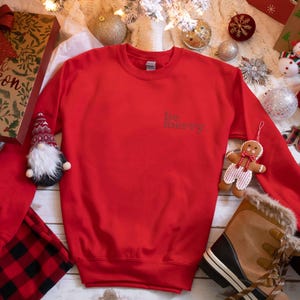 May include: Red Christmas sweatshirt with the text "Be Merry" in white on the front. The sweatshirt is surrounded by Christmas decorations, including a gnome, a gingerbread man, and a boot.