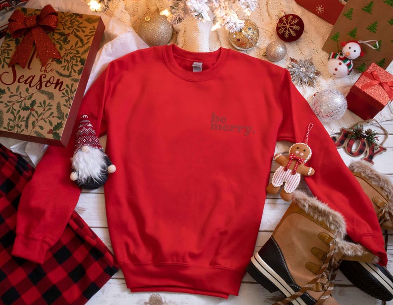 May include: Red Christmas sweatshirt with the text "Be Merry" in white on the front. The sweatshirt is surrounded by Christmas decorations, including a gnome, a gingerbread man, and a boot.