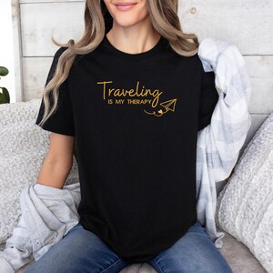 Może przedstawiać: Czarny t-shirt z napisem "Traveling is my therapy" w złotych literach i grafiką złotego papierowego samolotu.