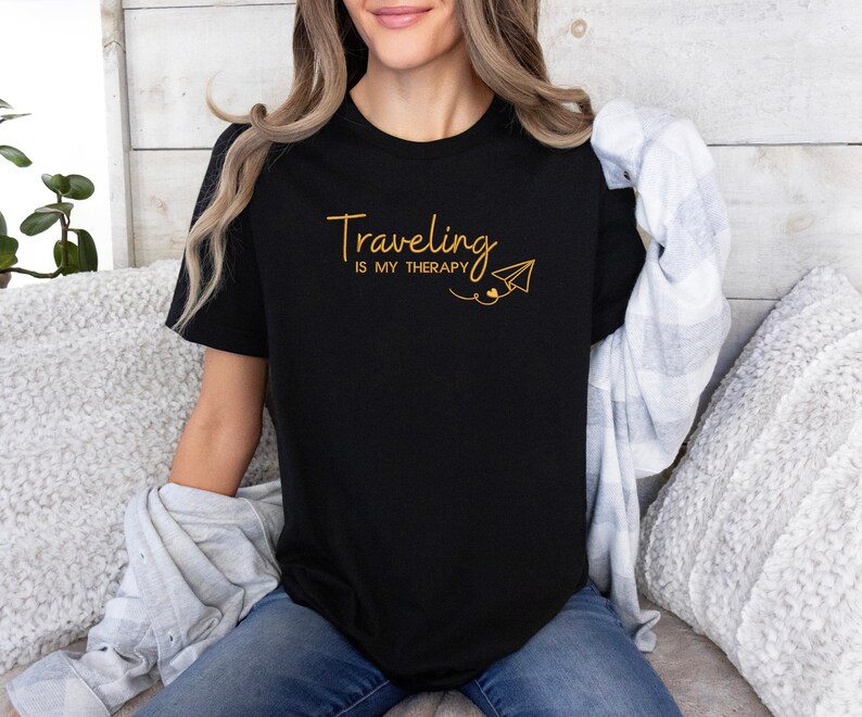 Może przedstawiać: Czarny t-shirt z napisem "Traveling is my therapy" w złotych literach i grafiką złotego papierowego samolotu.