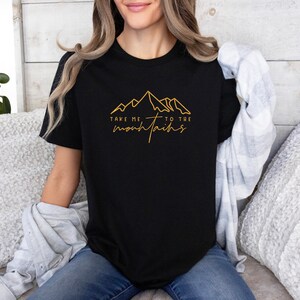 May include: A black t-shirt with a gold outline of mountains and the text "Take me to the mountains".
