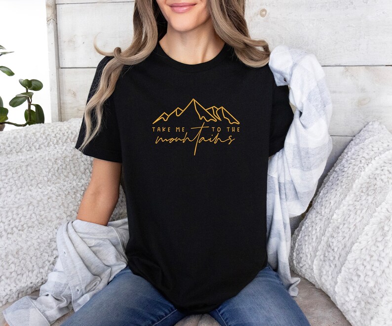 May include: A black t-shirt with a gold outline of mountains and the text "Take me to the mountains".