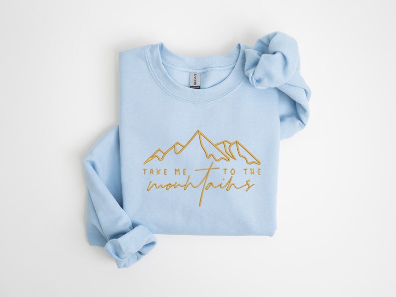 May include: Light blue sweatshirt with a gold outline of mountains and the text "Take me to the mountains".