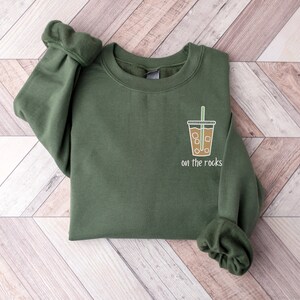 K&ouml;nnte beinhalten: Ein gr&uuml;nes Sweatshirt mit einem Aufdruck eines Kaffeebechers mit Eis und einem Strohhalm. Der Text "on the rocks" ist unter dem Becher gedruckt.