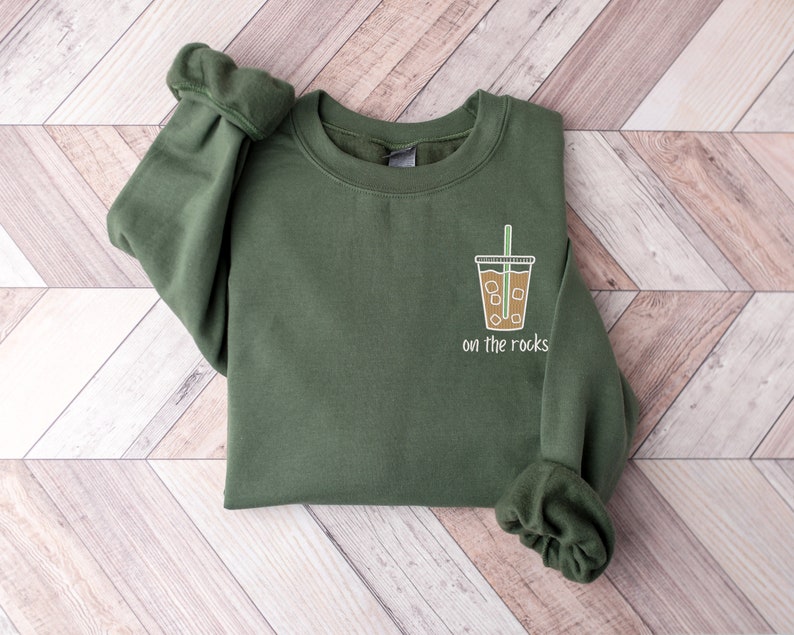 K&ouml;nnte beinhalten: Ein gr&uuml;nes Sweatshirt mit einem Aufdruck eines Kaffeebechers mit Eis und einem Strohhalm. Der Text "on the rocks" ist unter dem Becher gedruckt.