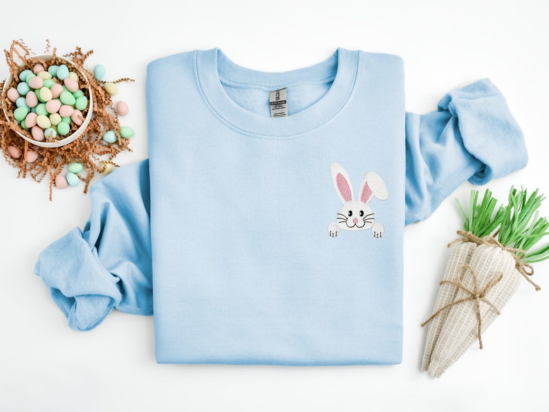 May include: A light blue sweatshirt with a white bunny rabbit face peeking out from the pocket. The bunny has pink ears and a black nose.
