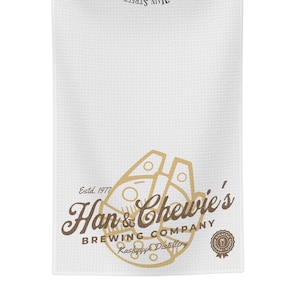 Może przedstawiać: Biały ręcznik kuchenny z brązowo-złotym logo, na którym widnieje napis "Han & Chewie's Brewing Company" wraz z tekstem "Estd. 1977" i "Kashyyyk Distillery". Logo zawiera stylizowane zdjęcie Sokoła Millennium.