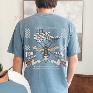 May include: A light blue t-shirt with a vintage-style graphic of a bee and the text "Fizzing Whizzbees". The graphic includes the text "Fruity Flavor!" and "Float for Minutes!" and a checkered pattern.