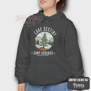May include: A dark gray hooded sweatshirt with a front pocket and a drawstring hood. The sweatshirt features a circular graphic with the words "Lake Destiny" and "Camp Grounds" with a nature scene. The text "Home of the Perfect Cast" is also included.