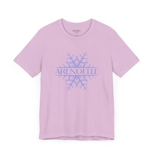 Op de afbeelding: Een lichtroze T-shirt met een blauw sneeuwvlokkenontwerp en het woord "ARENDELLE" erop gedrukt.