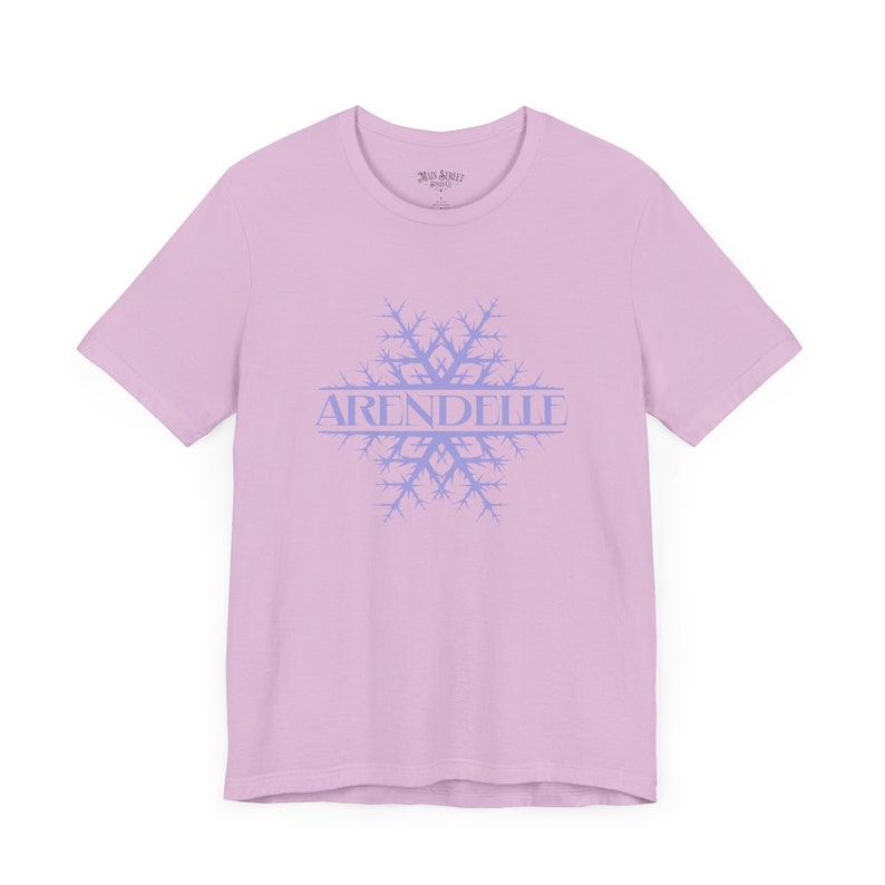 Op de afbeelding: Een lichtroze T-shirt met een blauw sneeuwvlokkenontwerp en het woord "ARENDELLE" erop gedrukt.