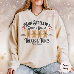 May include: Ivory-colored sweatshirt with the text "Main Street USA Dapper Bakery Treats & Tunes For The Holidays." The design includes gingerbread bear figures. The sweatshirt is a comfortable, casual style.