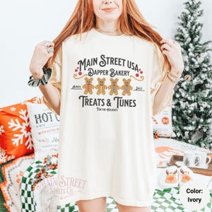 May include: A white t-shirt with a graphic design featuring gingerbread men and the text "Main Street USA Dapper Bakery Treats & Tunes For The Holidays".