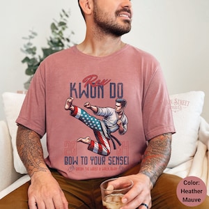 May include: A heather mauve t-shirt with a graphic of a person in a karate pose with an American flag design. The text "KWON DO" is above the graphic and "BOW TO YOUR SENSEI" is below the graphic. The text "20 64" is on either side of the graphic.