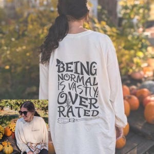 May include: A white long-sleeved shirt with a black graphic print that says "Being normal is vastly overrated" with a witch's hat and broom.