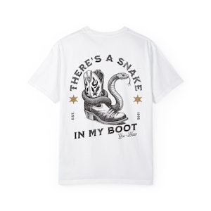May include: White t-shirt with a black and white graphic print of a snake in a cowboy boot. The graphic includes the text "There's a Snake in My Boot" and "Yee-Haw".