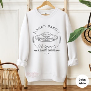 May include: White sweatshirt with a black and white graphic design of a bakery logo. The logo says "Tiana's Bakery" and features a plate of beignets with the text "Beignets & Baked Goods" and "Dreams do come true in New Orleans".