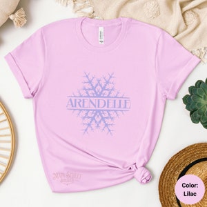 Op de afbeelding: Een lila t-shirt met een sneeuwvlokdesign en het woord "Arendelle" in de sneeuwvlok. Het shirt is gemaakt van een zacht, comfortabel materiaal.