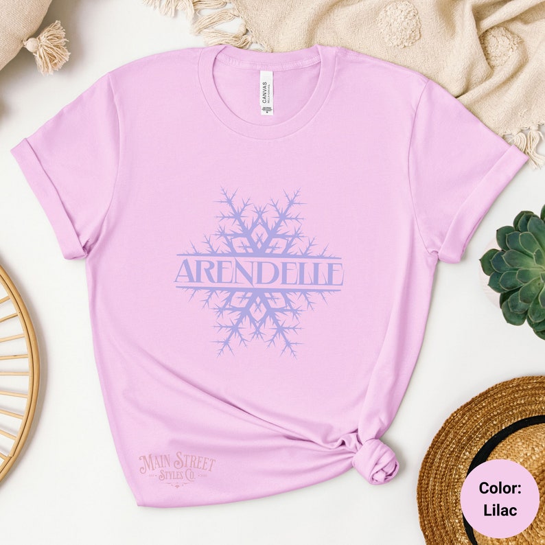 Op de afbeelding: Een lila t-shirt met een sneeuwvlokdesign en het woord "Arendelle" in de sneeuwvlok. Het shirt is gemaakt van een zacht, comfortabel materiaal.