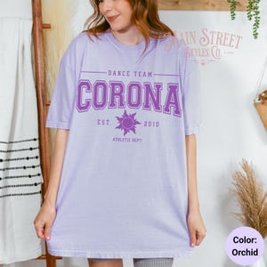 May include: Orchid-colored t-shirt with the text "CORONA" in a collegiate font, above the words "DANCE TEAM" and below "EST. 2010 ATHLETIC DEPT." A sun graphic is between "EST." and "2010". The shirt is a loose fit.