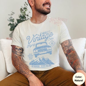 May include: A white t-shirt with a blue graphic design of a vintage car flying over mountains. The graphic includes the text "WEASLEY'S Vintage Cars Est. 1992 Flying For the adventurous witch or wizard The Burrow - Devon England".