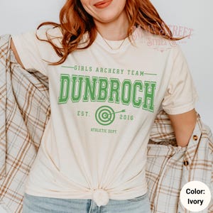 May include: Ivory-colored t-shirt with the text "GIRLS ARCHERY TEAM DUNBROCH EST. 2016 ATHLETIC DEPT." in green. The shirt features a bullseye graphic with an arrow. The model is wearing a plaid shirt.