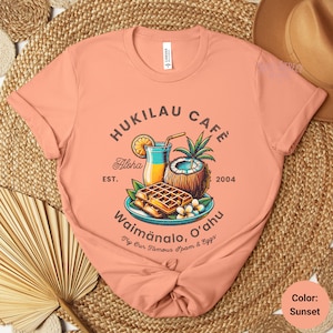 May include: A peach-colored t-shirt with a vintage-style logo for "Hukilau Cafe" featuring a tropical drink, a coconut, waffles, and the text "Aloha", "Est. 2004", "Waimanalo, O'ahu", and "Try Our Famous Spam & Eggs".