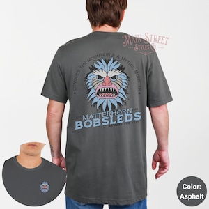 May include: A dark gray t-shirt with a graphic of a blue yeti-like creature with the text "Conquer the Mountain & a Mythic Monster" and "Est. Matterhorn Bobsleds Beware the Yeti".