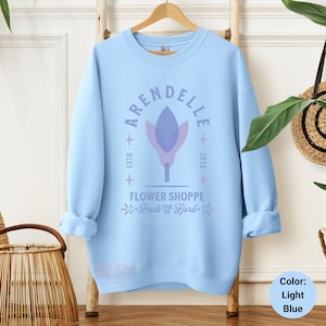 May include: Light blue sweatshirt with the text "ARENDELLE FLOWER SHOPPE Fresh from the Fjord" and a stylized flower graphic. The sweatshirt is hanging on a wooden hanger against a white wall.