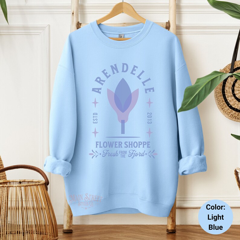 May include: Light blue sweatshirt with the text "ARENDELLE FLOWER SHOPPE Fresh from the Fjord" and a stylized flower graphic. The sweatshirt is hanging on a wooden hanger against a white wall.