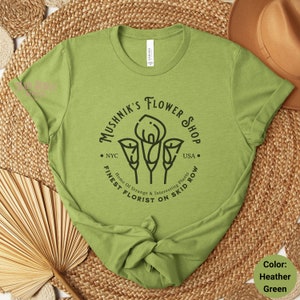 May include: A green t-shirt with a black graphic of a flower shop logo. The logo says "Mushnik's Flower Shop" with the text "NYC" and "USA" on either side of the flower graphic. The text below the flower graphic says "Finest Florist on Skid Row" and "Home of Strange & Interesting Plants".