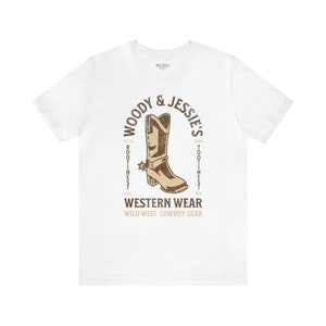 Może przedstawiać: Biała koszulka z brązowym nadrukiem kowbojskiego buta z tekstem "Woody & Jessie's" w k&oacute;łku wok&oacute;ł buta. Tekst "Western Wear" znajduje się pod butem, a tekst "Wild West Cowboy Gear" pod nim.