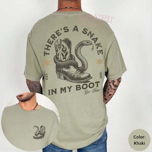 May include: A khaki green t-shirt with a graphic print of a cowboy boot with a snake wrapped around it. The text on the shirt reads "There's a Snake in My Boot Yee-Haw" with the dates "EST. 1995".