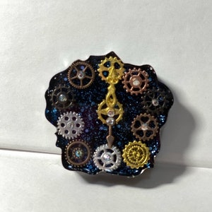 May include: A steampunk-style brooch with a dark blue, glittery backdrop. The brooch showcases an assortment of metal gears in gold, silver, and bronze hues, some with clear crystal embellishments. The brooch has a distinctive, irregular form.